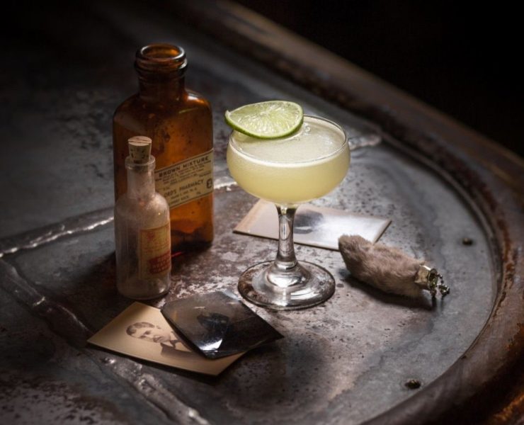 A cocktail glass with a lime slice on a metal surface, surrounded by two vintage bottles, old photographs, and a small cloth bundle.