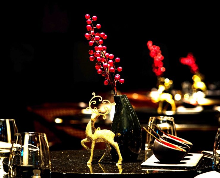 A dimly lit dining table adorned with glasses, bowls, and a centerpiece featuring a gold reindeer figurine beside a vase with red berries.