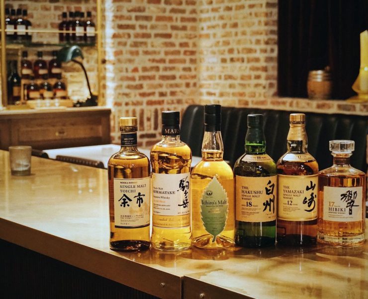 Six bottles of Japanese whisky lined up on a wooden bar counter in a warmly lit room with exposed brick walls.