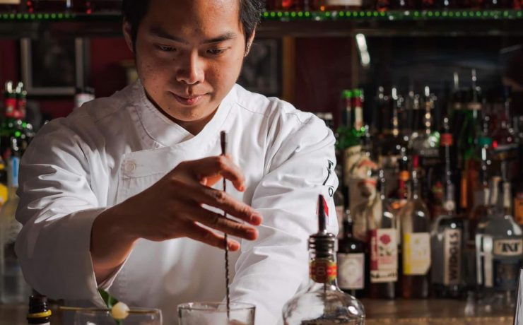 Bartender in white coat stirs an iced drink with a spoon at a bar full of various liquor bottles.