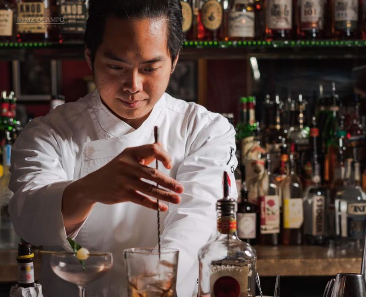Bartender in white coat stirs an iced drink with a spoon at a bar full of various liquor bottles.