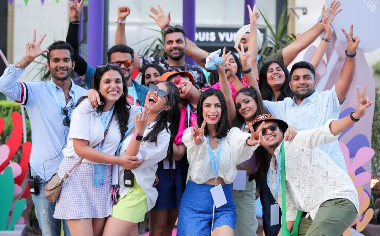 A group of people smiling and posing for a photo outdoors. Some are making peace signs and wearing casual, festive clothing. Colorful decorations and a building are in the background.