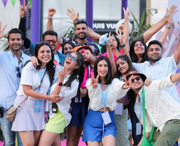A group of people smiling and posing for a photo outdoors. Some are making peace signs and wearing casual, festive clothing. Colorful decorations and a building are in the background.