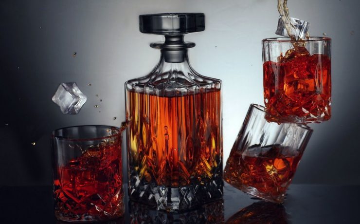 A glass decanter filled with whiskey is surrounded by three whiskey glasses, one tipped over, scattering ice and liquid.