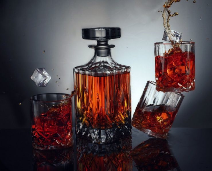 A glass decanter filled with whiskey is surrounded by three whiskey glasses, one tipped over, scattering ice and liquid.