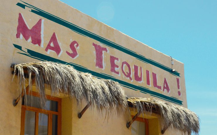 Building facade with a sign saying "MAS TEQUILA!" in red letters. Straw awnings are above the windows. Sky is clear blue.