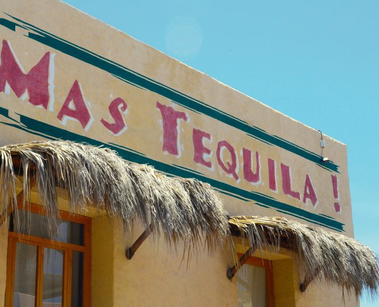 Building facade with a sign saying "MAS TEQUILA!" in red letters. Straw awnings are above the windows. Sky is clear blue.