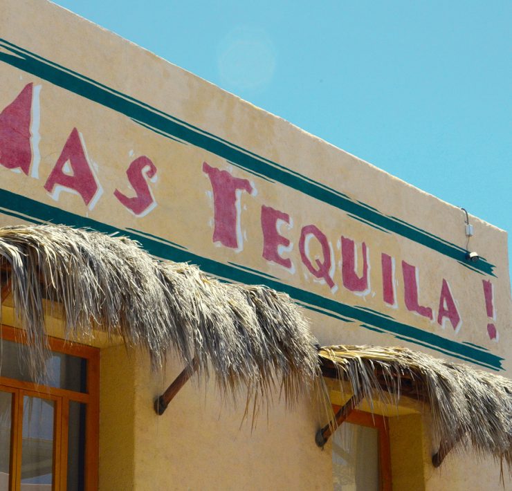 Building facade with a sign saying "MAS TEQUILA!" in red letters. Straw awnings are above the windows. Sky is clear blue.