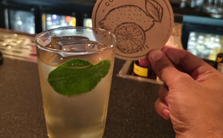 A person holds a coaster with a citrus design beside a glass of light-colored beverage garnished with a lime leaf on a bar counter.