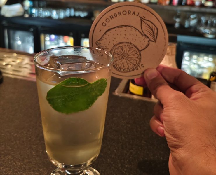 A person holds a coaster with a citrus design beside a glass of light-colored beverage garnished with a lime leaf on a bar counter.