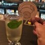 A person holds a coaster with a citrus design beside a glass of light-colored beverage garnished with a lime leaf on a bar counter.